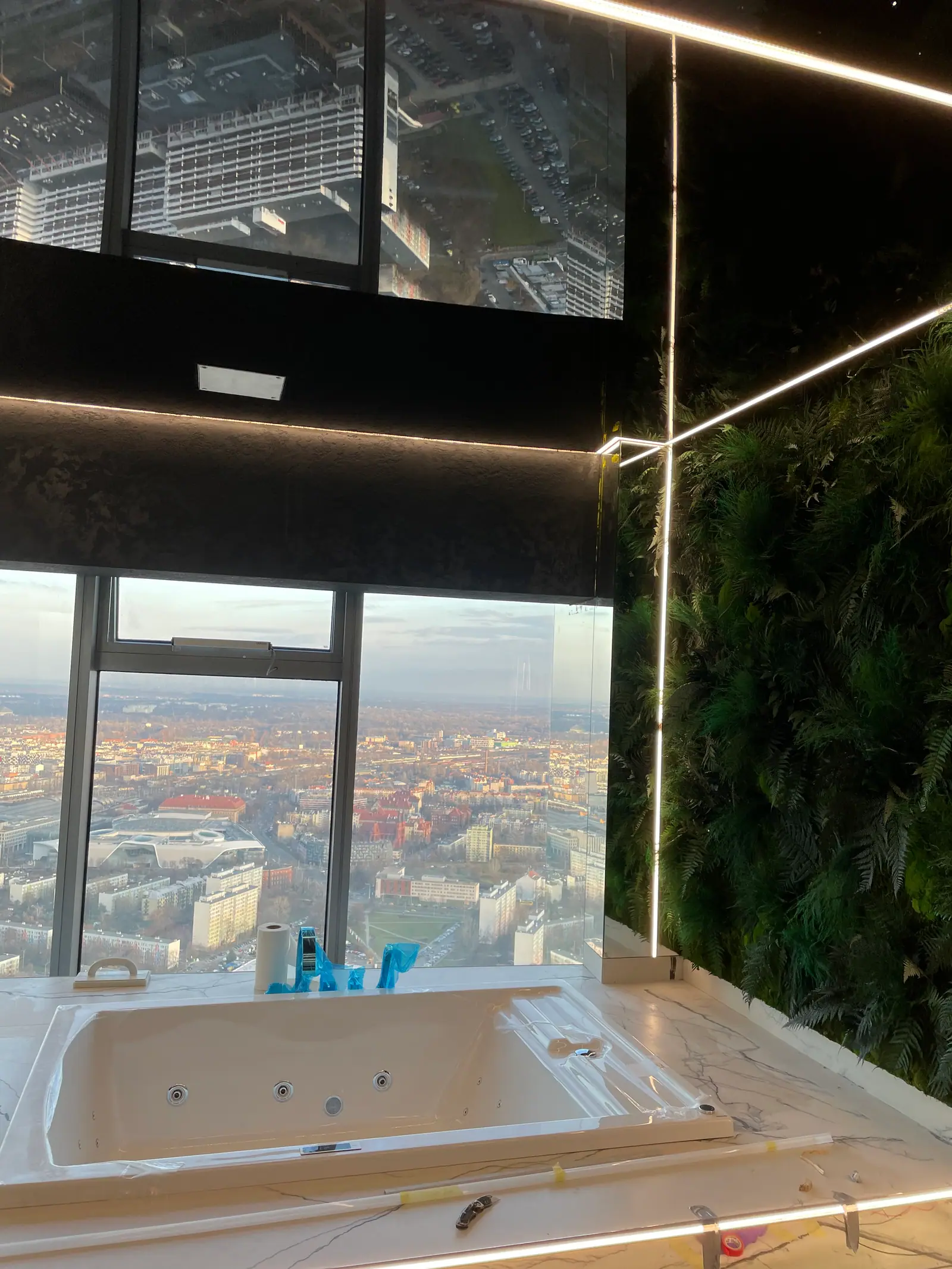 A whirlpool bathtub by a window with a city view, a plant wall and LED strips beside it, tools lying on the surround.