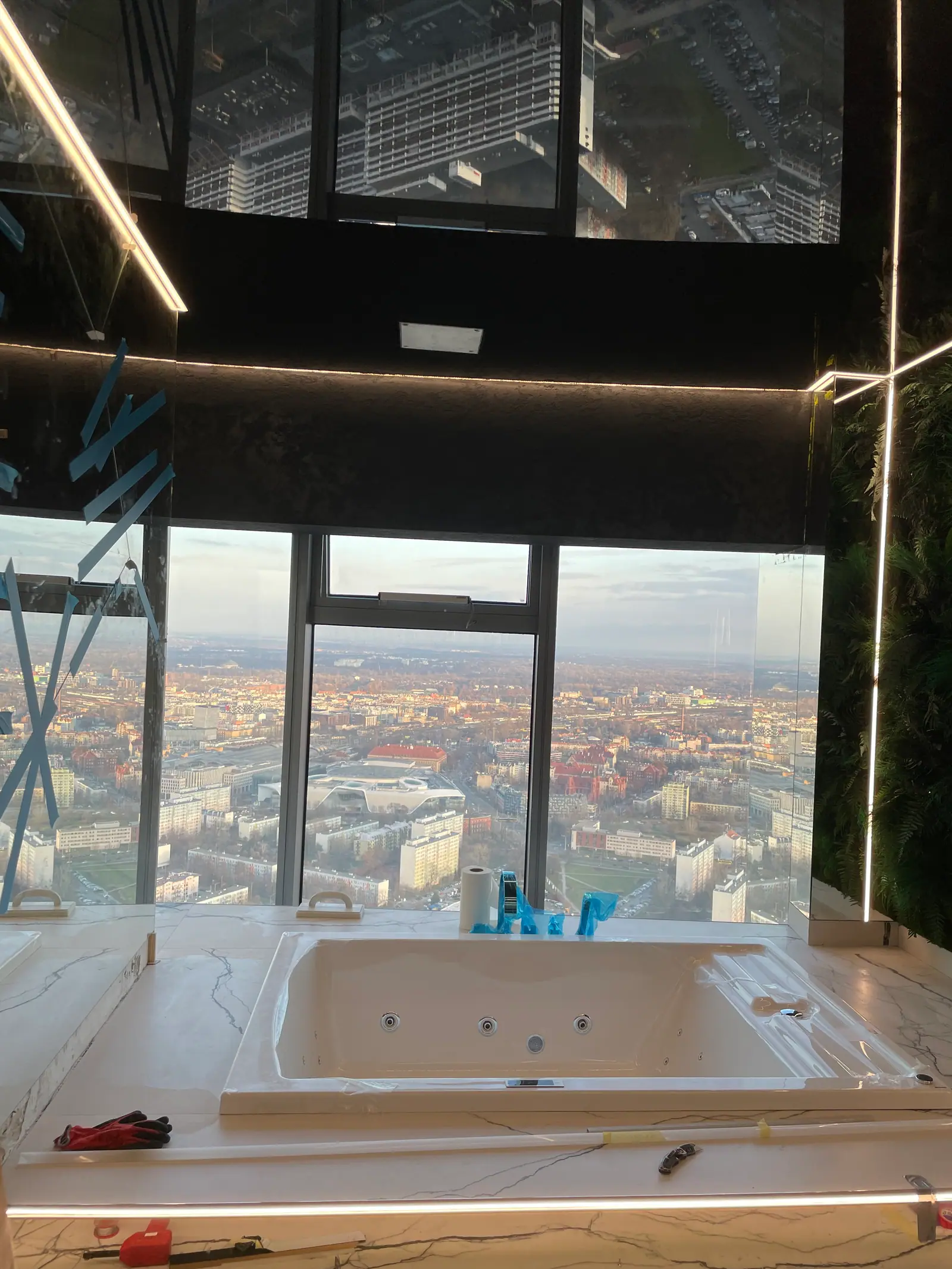 A bathtub in a light surround by windows with a city skyline, a mirrored ceiling, tape and tools during installation.