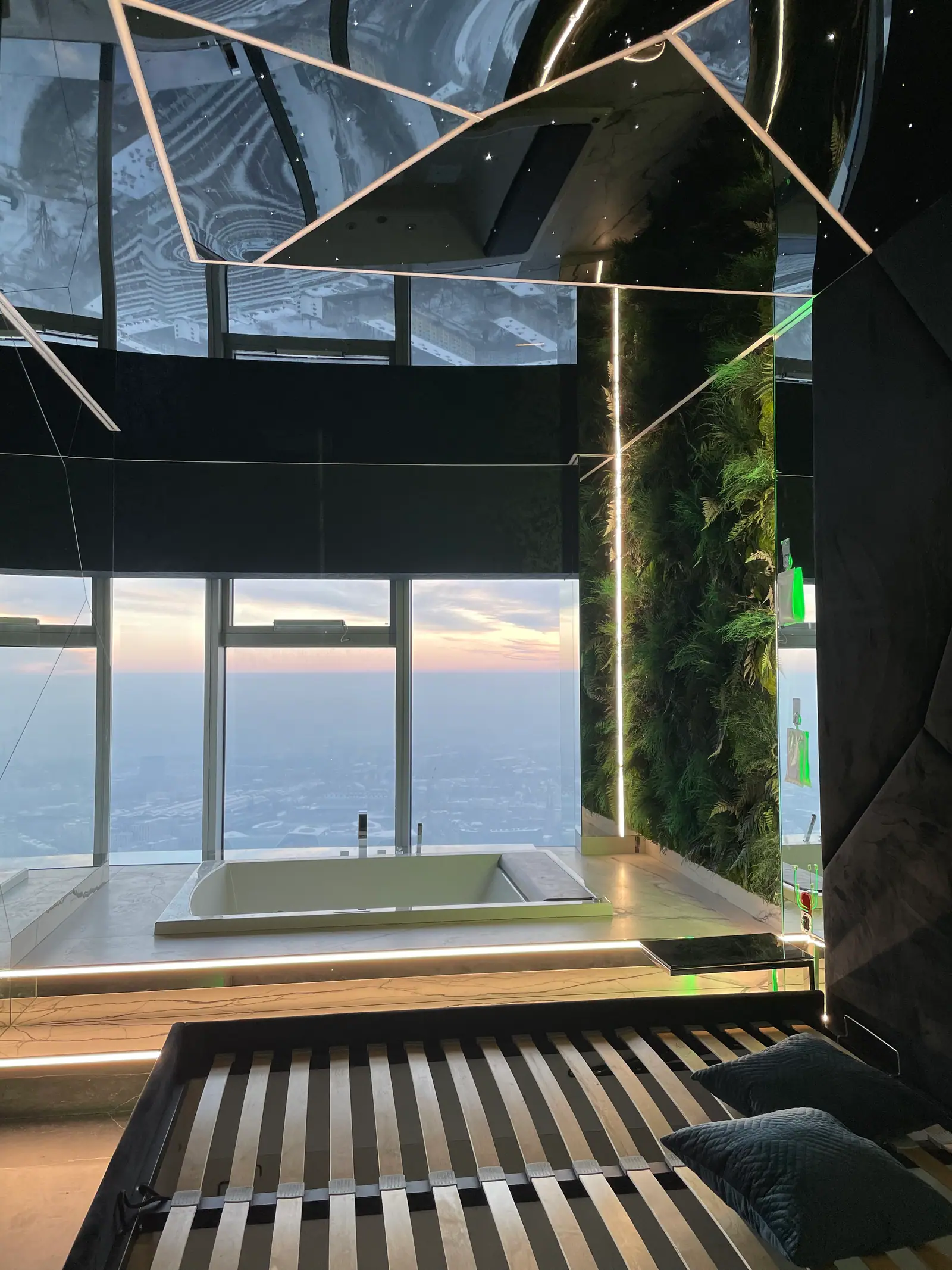 A bed frame, a bathtub on a platform by the glazed wall, a green wall and a mirrored ceiling with LED strips, with the sunset outside.