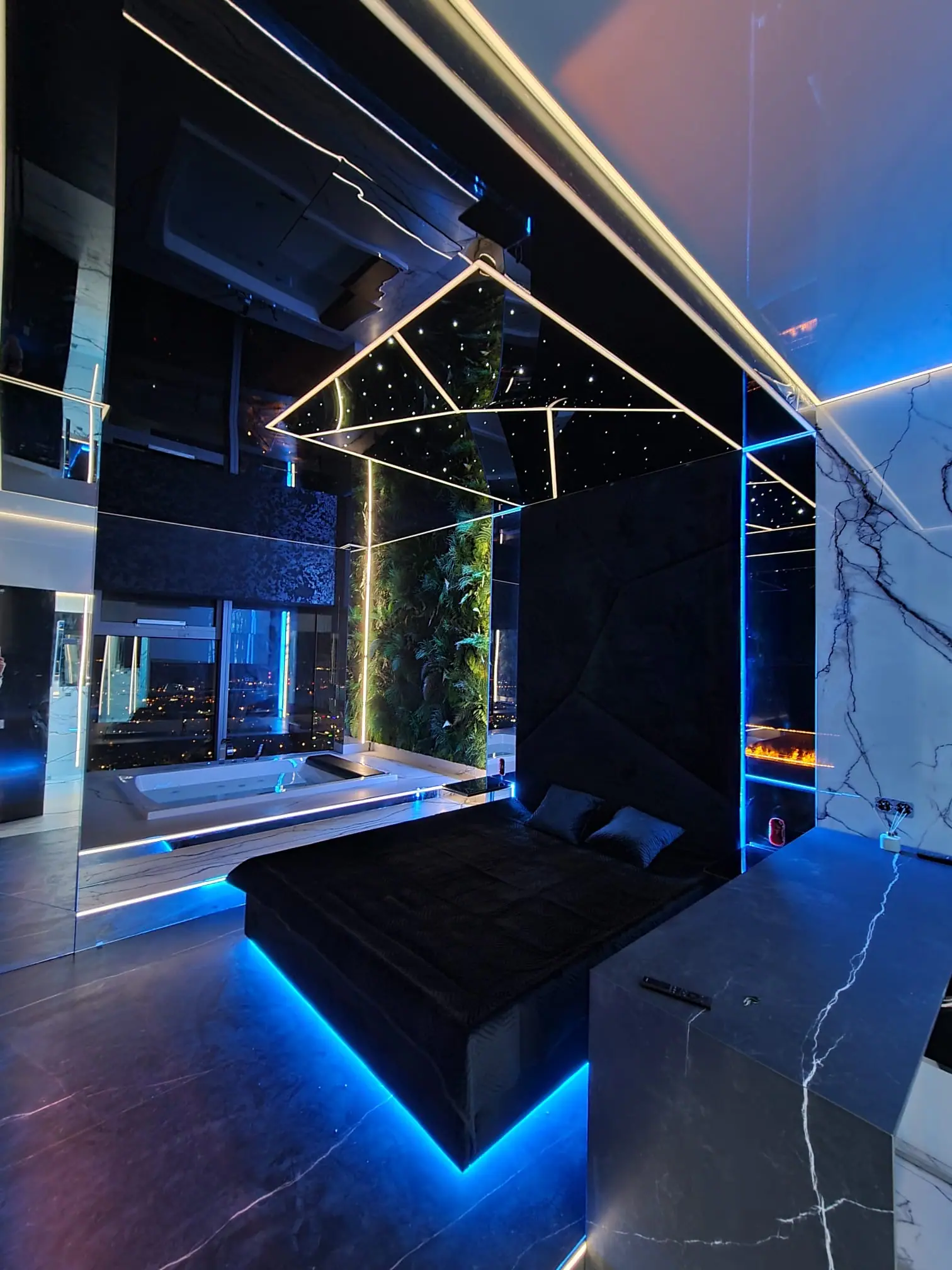 A dark interior in the evening: a backlit bed, a ceiling with points of light, a green wall and a bathtub by the window with a view of the city.