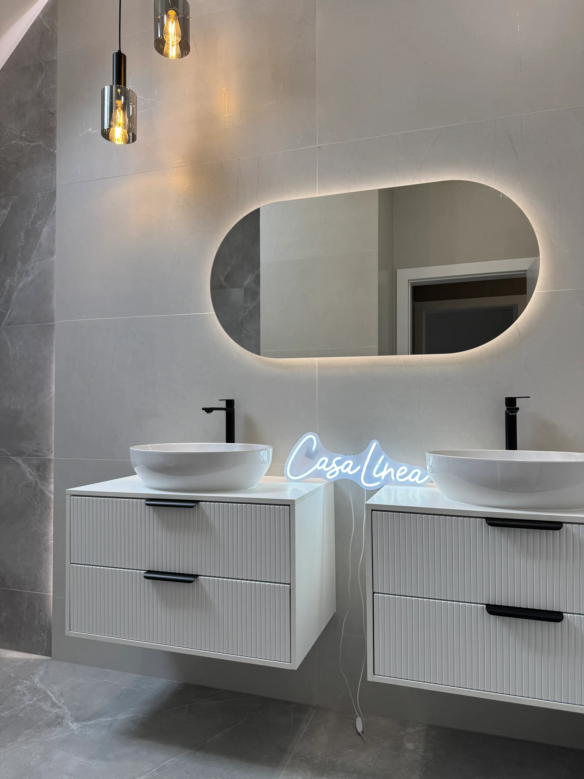 Bathroom with two basins on white vanity units, an oval backlit mirror, grey tiles and a Casa Linea neon sign.
