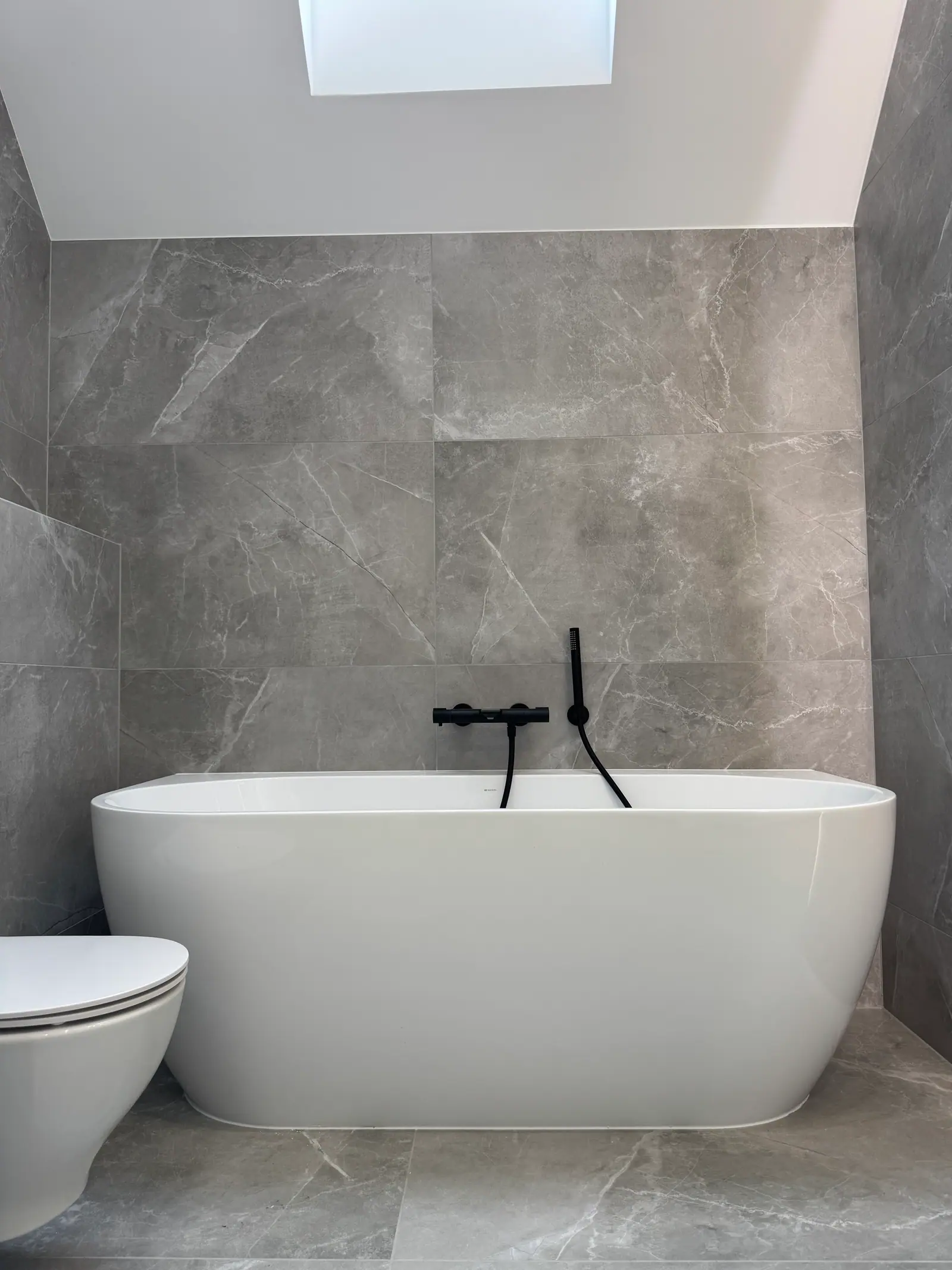 A white freestanding bathtub with a black wall-mounted tap under a sloping ceiling with a roof window, walls in grey tiles.