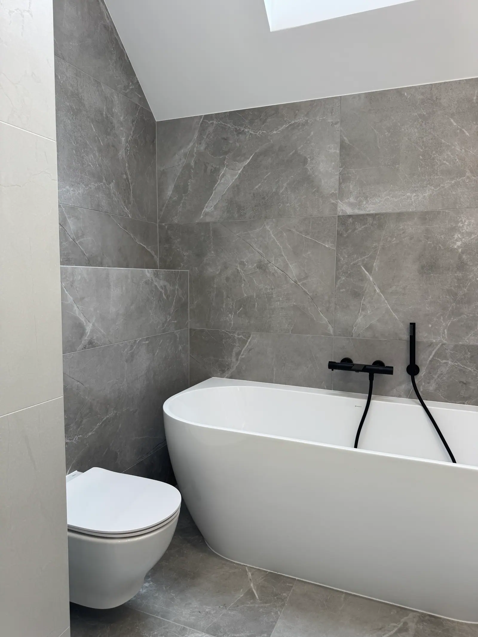 A freestanding bathtub and a wall-hung toilet in a bathroom with grey tiles, seen from the side.