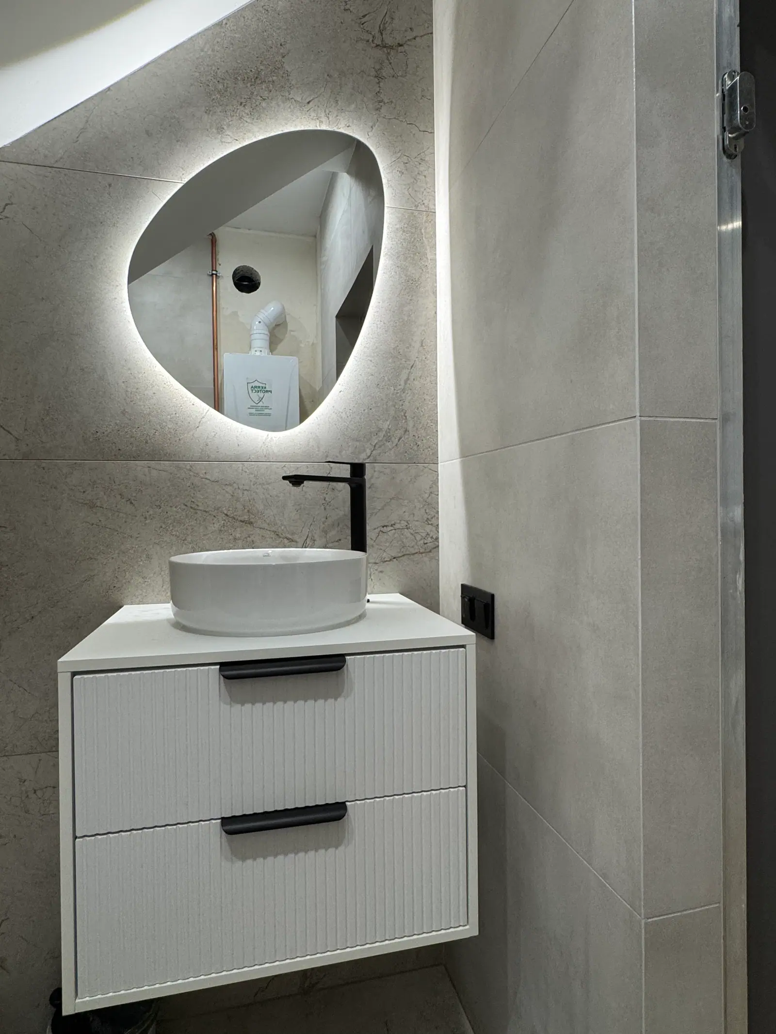 A small bathroom with a round basin on a white vanity unit, a black tap and a backlit mirror with an irregular shape.