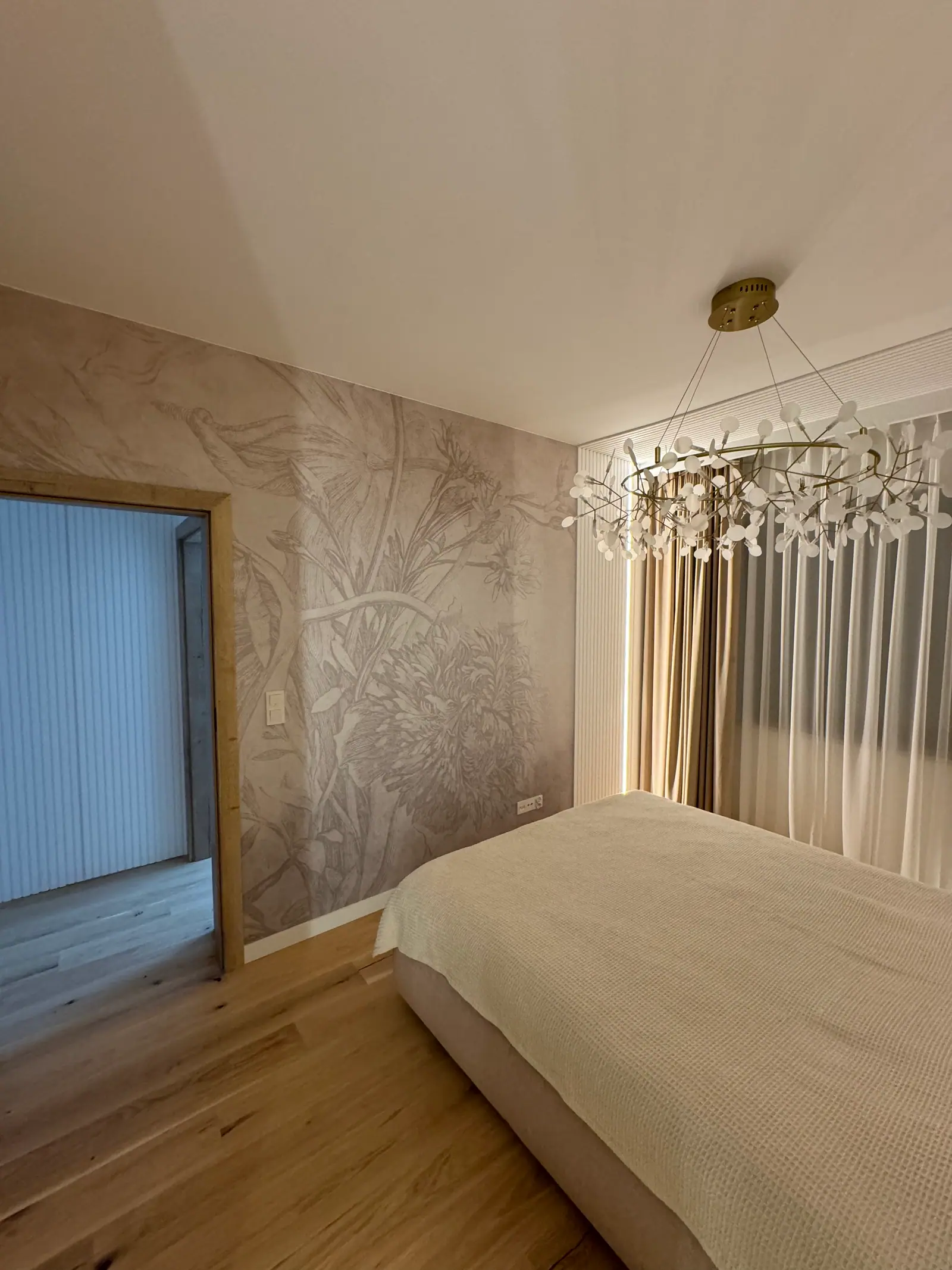Bedroom with botanical wallpaper, a chandelier of glass shades, curtains and a wooden floor.