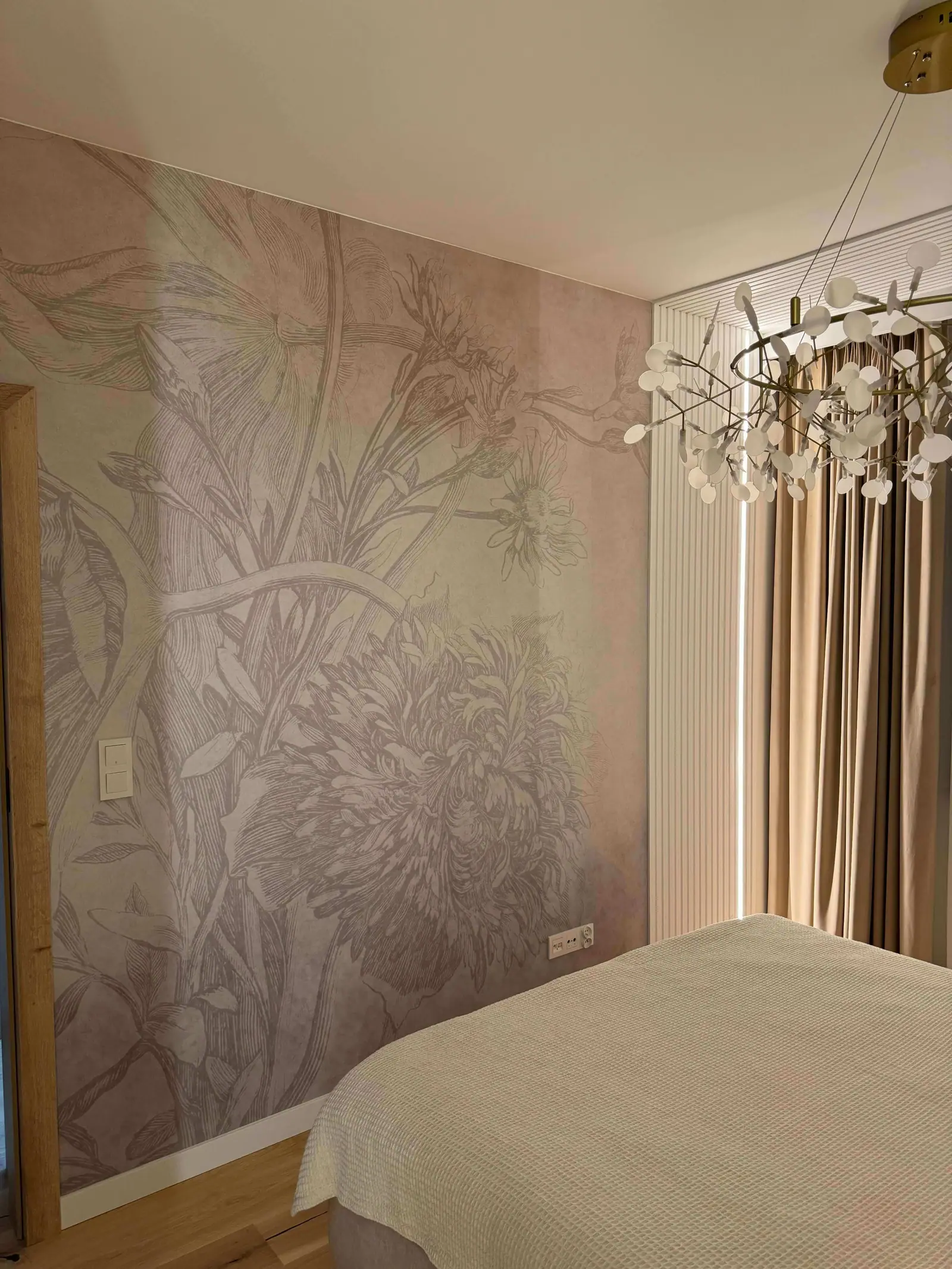 The bedroom from another angle: the bed, botanical wallpaper, a chandelier of glass shades, curtains and an open doorway.