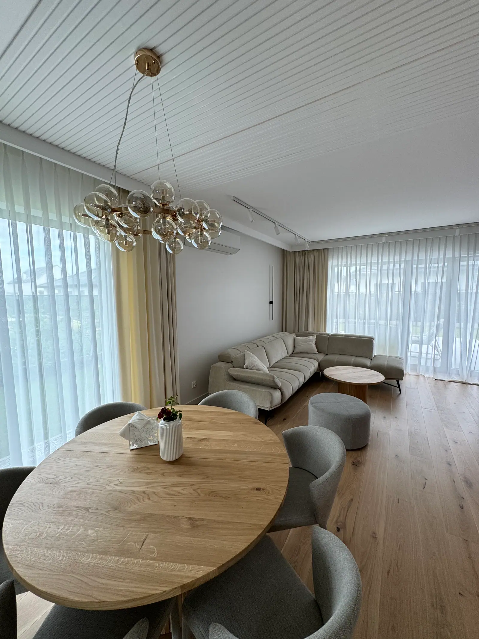 A round wooden table with grey chairs under a chandelier of glass globes, with the living room behind.