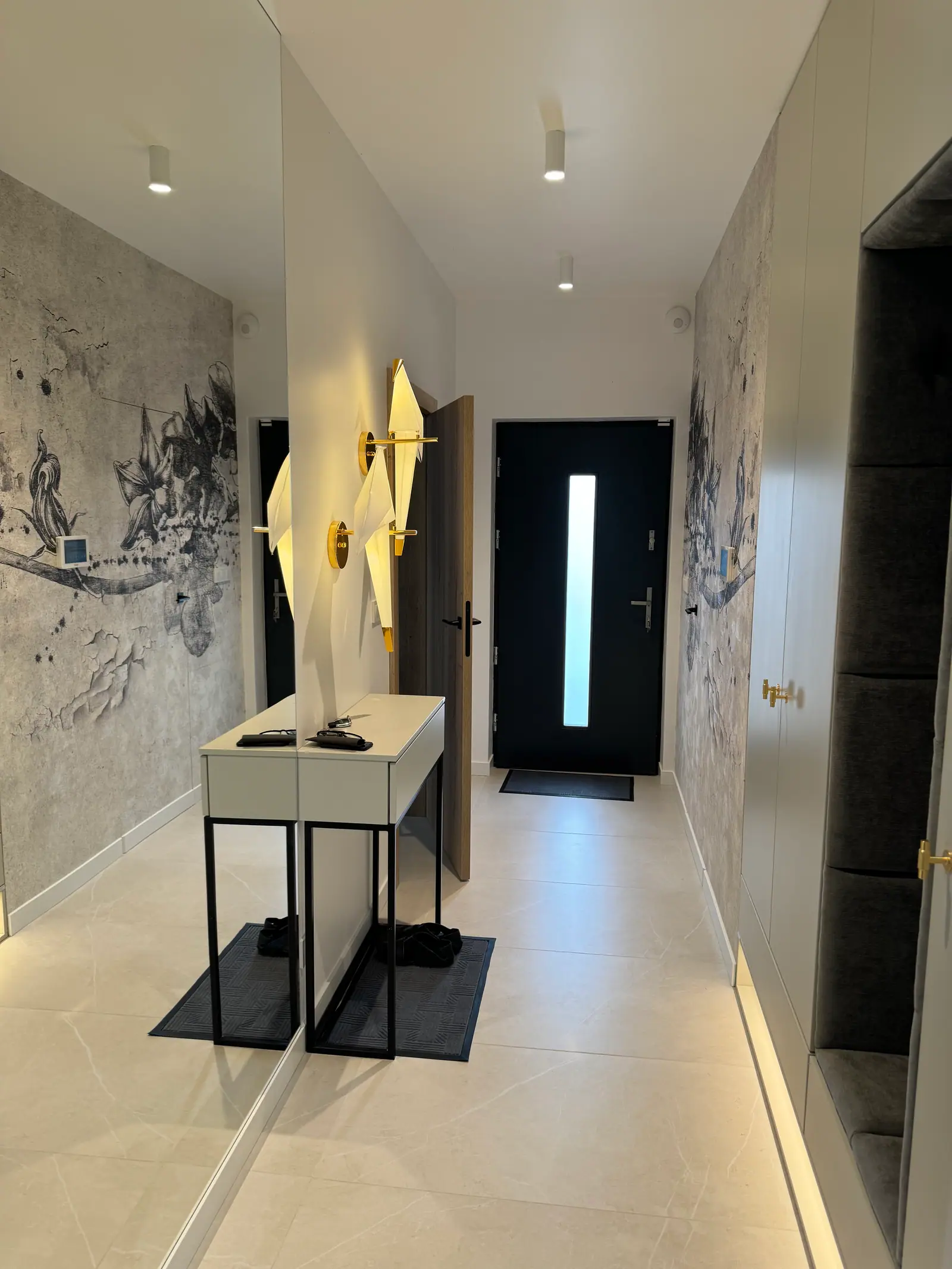 Hallway with dark-patterned wallpaper, a console table, gold wall lights, a mirror and a dark front door.