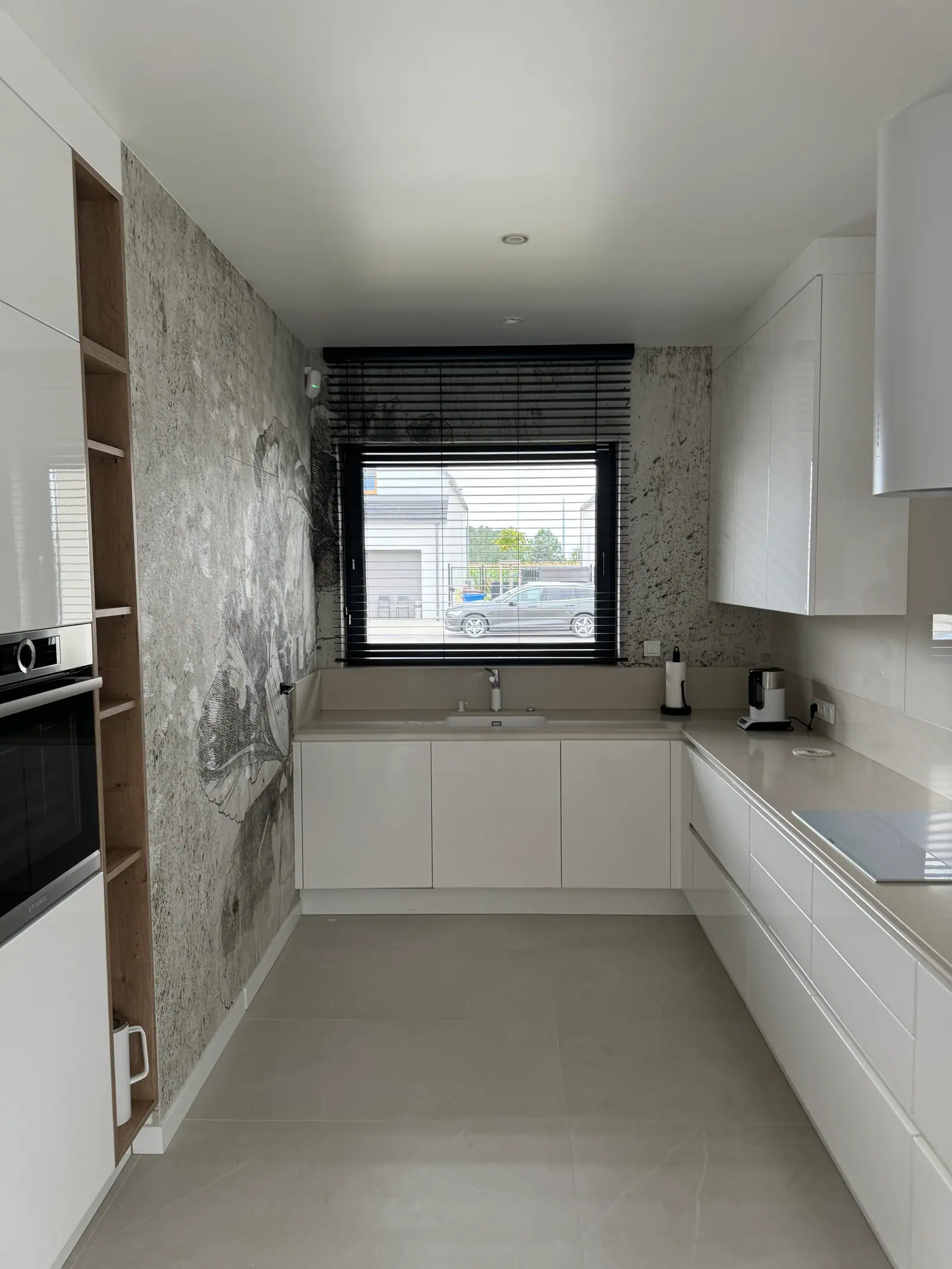 Kitchen with white L-shaped cabinets, a light worktop, a window with a blind and wallpaper with a dark pattern.