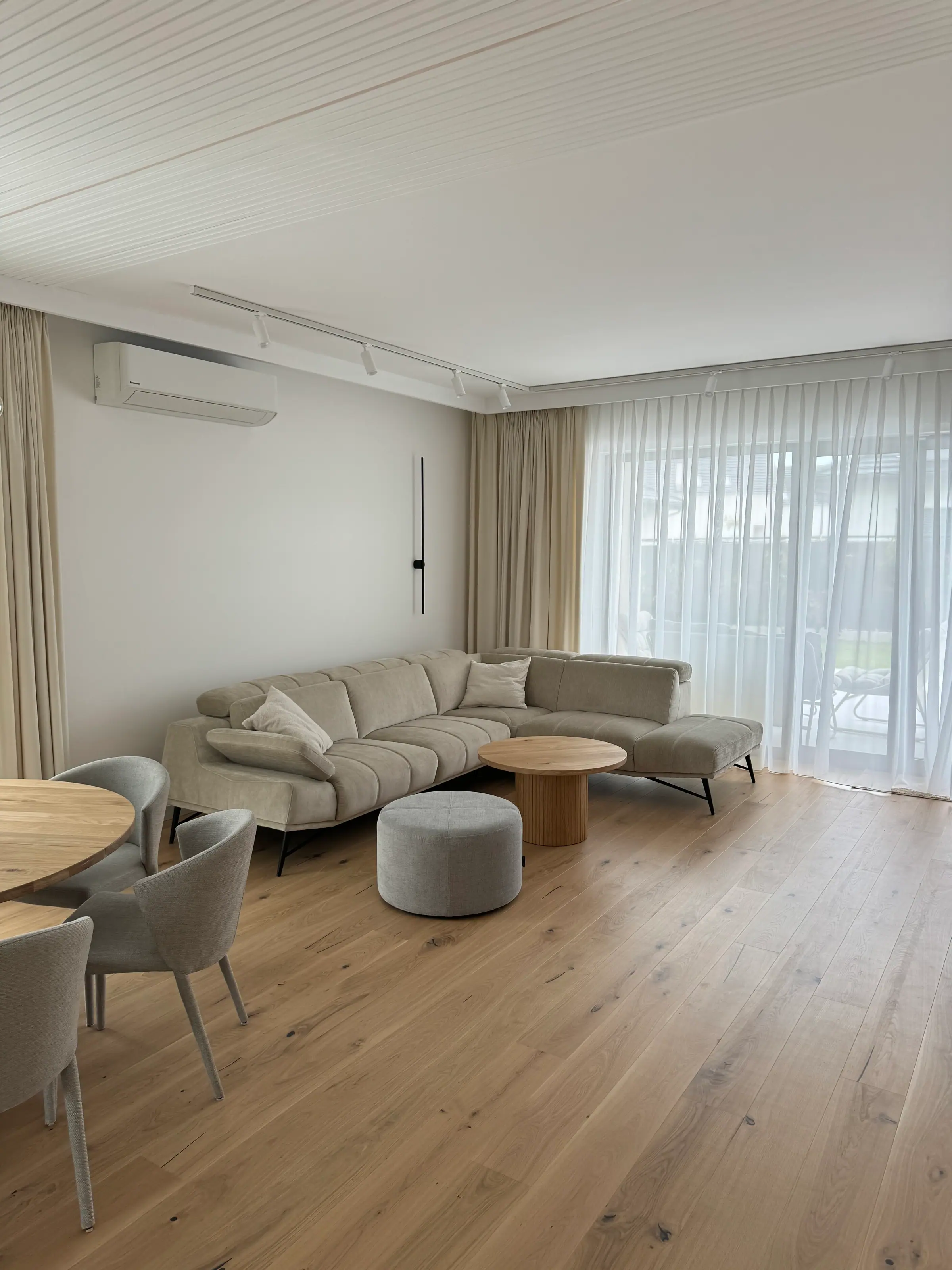Living room with a light corner sofa, a round coffee table and a pouffe, a wooden floor and windows with sheer curtains.