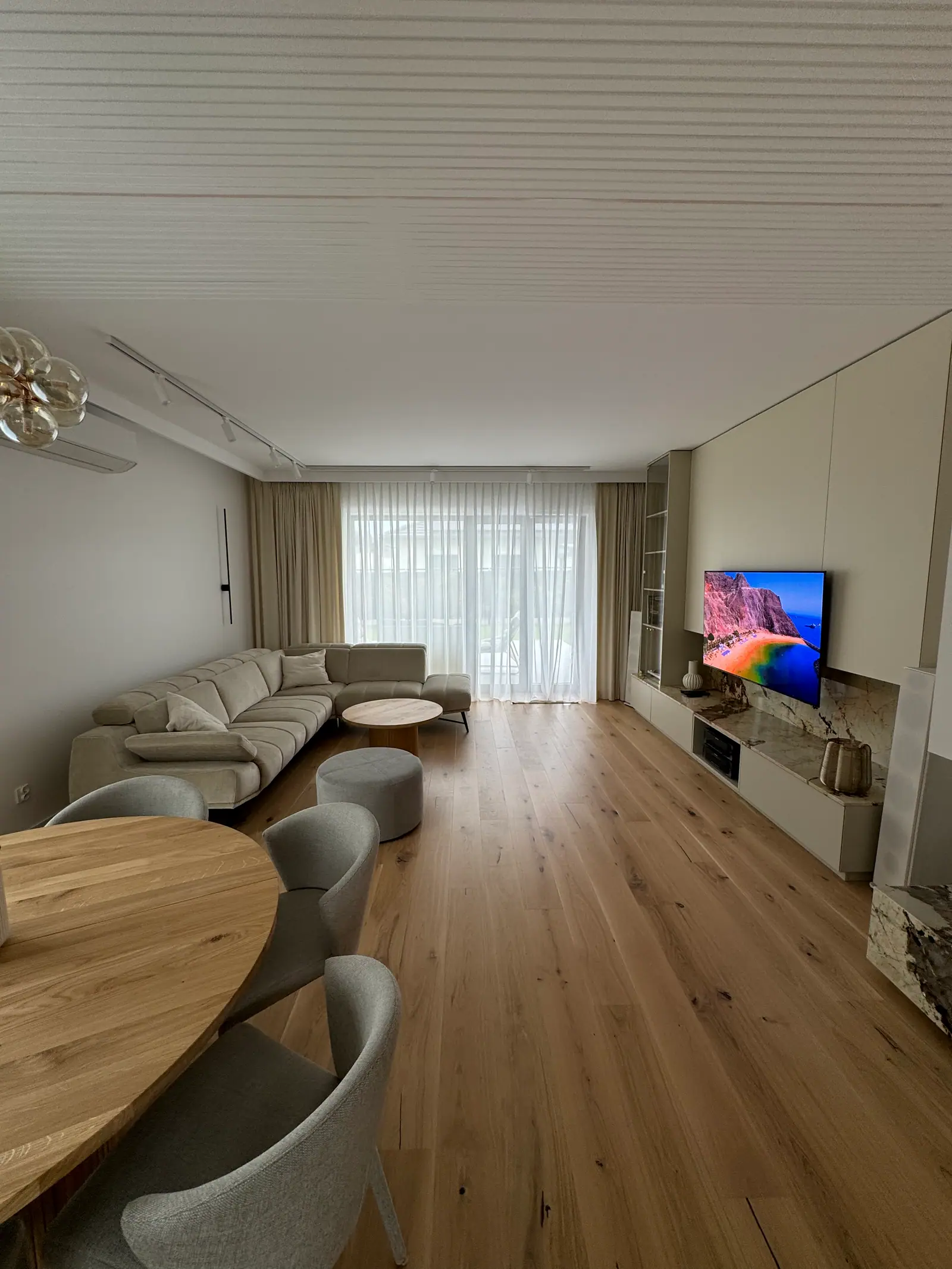 The living room seen from the table: corner sofa, a built-in wall with a television and a ceiling of white slats.