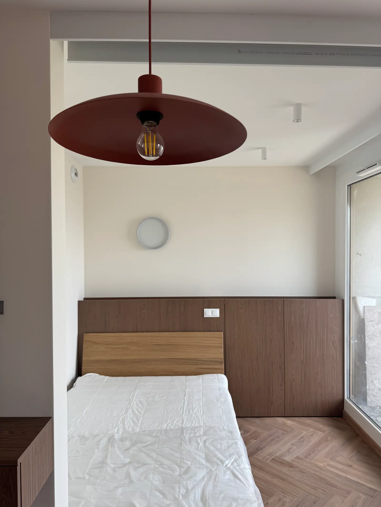 Bedroom with a bed against a wall lined with wood-coloured panels, a wall light and a red pendant lamp.
