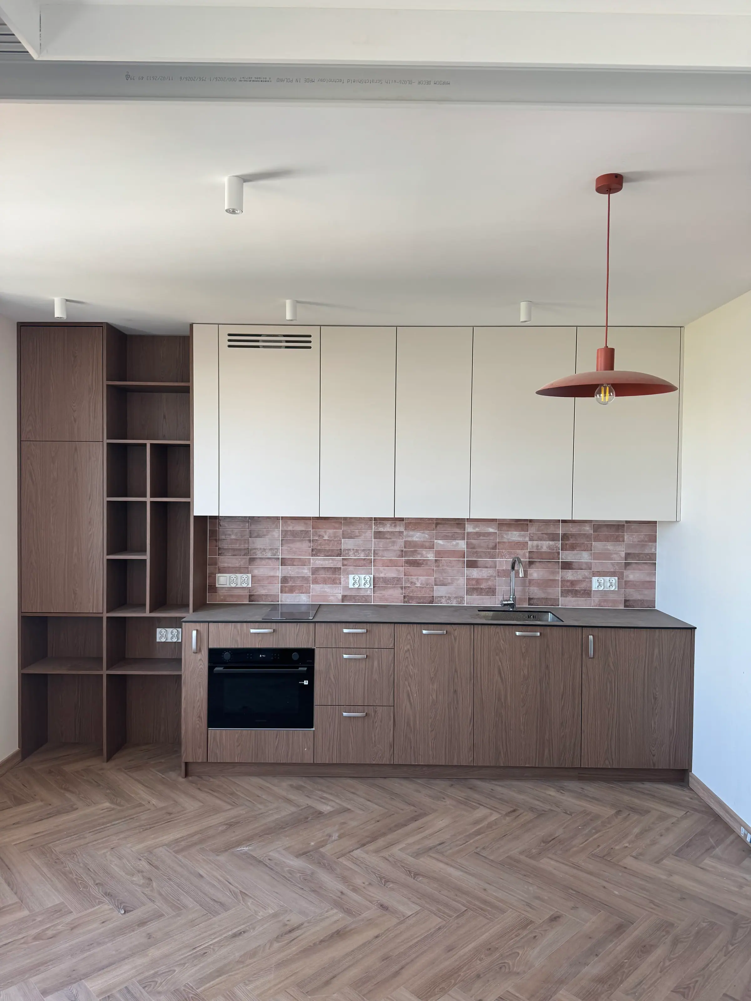 Kitchen with wood-coloured cabinets and cream wall units, brick-toned tiles, a herringbone floor and a red pendant lamp.