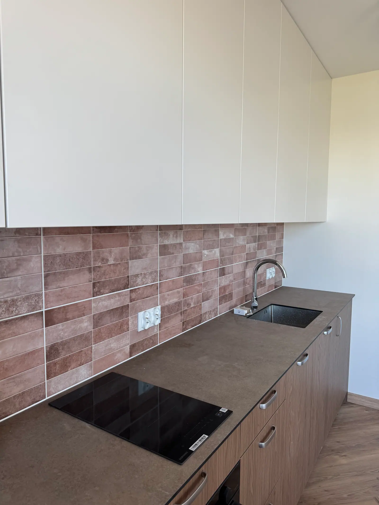 A kitchen worktop with a hob and a sink, brick-toned tiles and cream wall units.